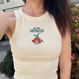 May include: Cream-colored tank top with the words "The Little Mermaid" in teal and a cartoon image of Ariel. The top is ribbed and has a round neckline.