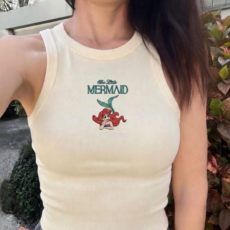 May include: Cream-colored tank top with the words "The Little Mermaid" in teal and a cartoon image of Ariel. The top is ribbed and has a round neckline.