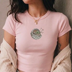 K&ouml;nnte beinhalten: Hellrosa T-Shirt mit einer Cartoon-Illustration eines gr&uuml;nen Alien-Charakters in einer Kapsel, mit Noten. Das Design befindet sich auf der Vorderseite des T-Shirts. Das T-Shirt hat kurze &Auml;rmel und einen Rundhalsausschnitt.
