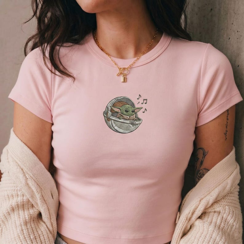 K&ouml;nnte beinhalten: Hellrosa T-Shirt mit einer Cartoon-Illustration eines gr&uuml;nen Alien-Charakters in einer Kapsel, mit Noten. Das Design befindet sich auf der Vorderseite des T-Shirts. Das T-Shirt hat kurze &Auml;rmel und einen Rundhalsausschnitt.