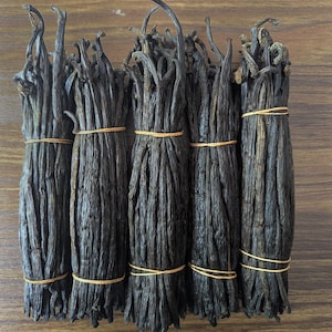 Peut inclure: Cinq bouquets de gousses de vanille brun foncé, attachés avec des élastiques jaunes, sont disposés sur une surface en bois. Les gousses ont un aspect texturé, sur un fond de grain de bois brun clair.