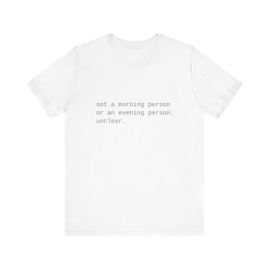 以下が含まれることがあります： 「not a morning person or an evening person. unclear.」というテキストがプリントされた白いTシャツ。シンプルなサンセリフ体のフォントで書かれています。クルーネックのベーシックなスタイルです。