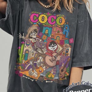 May include: A charcoal gray oversized t-shirt featuring a colorful graphic print from the movie Coco, depicting a skeleton character and a boy playing a guitar in front of a Mexican-style building. The text Comfort Colors Pepper is visible.