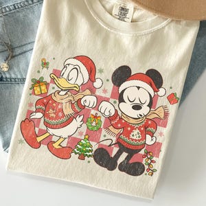 May include: A cream-coloured t-shirt featuring a graphic of Donald Duck and Mickey Mouse wearing red Santa hats and patterned Christmas jumpers. The design includes a pink checkered background, a small Christmas tree, a wreath, and gifts.