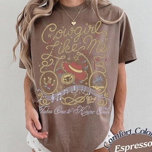 Op de afbeelding: Een bruin t-shirt met korte mouwen en een grafische print met de tekst Cowgirl Like Me en Takes One to Know One. Het ontwerp bevat een rode cowboyhoed, een lasso, muzieknoten en de zin Eyes full of Stars in wit en geel.
