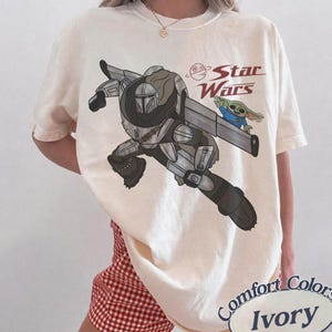 Pu&ograve; includere: T-shirt color avorio con una grafica di The Mandalorian e Grogu di Star Wars. Il design include il logo di Star Wars in rosso. La maglietta &egrave; etichettata "Comfort Colors" e "Ivory".
