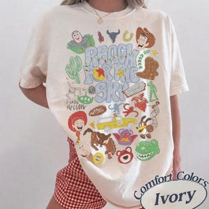 Peut inclure: Un t-shirt ivoire avec une impression graphique color&eacute;e portant le texte "REACH FOR THE SKY" et divers personnages de dessins anim&eacute;s et ic&ocirc;nes, dont un cow-boy, un ranger de l'espace, un dinosaure, un extraterrestre et un camion jaune.