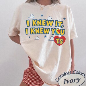K&ouml;nnte beinhalten: Ein elfenbeinfarbenes Kurzarm-T-Shirt mit dem blau-gelben Text I KNEW IT, I KNEW YOU und einem roten Herz mit den Buchstaben TS. Das Design enth&auml;lt kleine blaue Sterne und wei&szlig;e Wolkenumrisse, wobei eine Ballgrafik den Buchstaben O ersetzt.