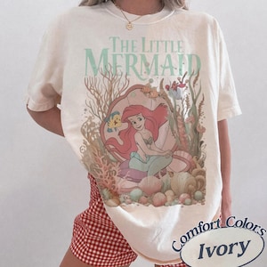 Pu&ograve; includere: Una t-shirt a maniche corte color avorio con una stampa grafica de La Sirenetta che raffigura Ariel, Flounder ed elementi di corallo sottomarino. Il design include il testo The Little Mermaid in un carattere verde chiaro. Girocollo.