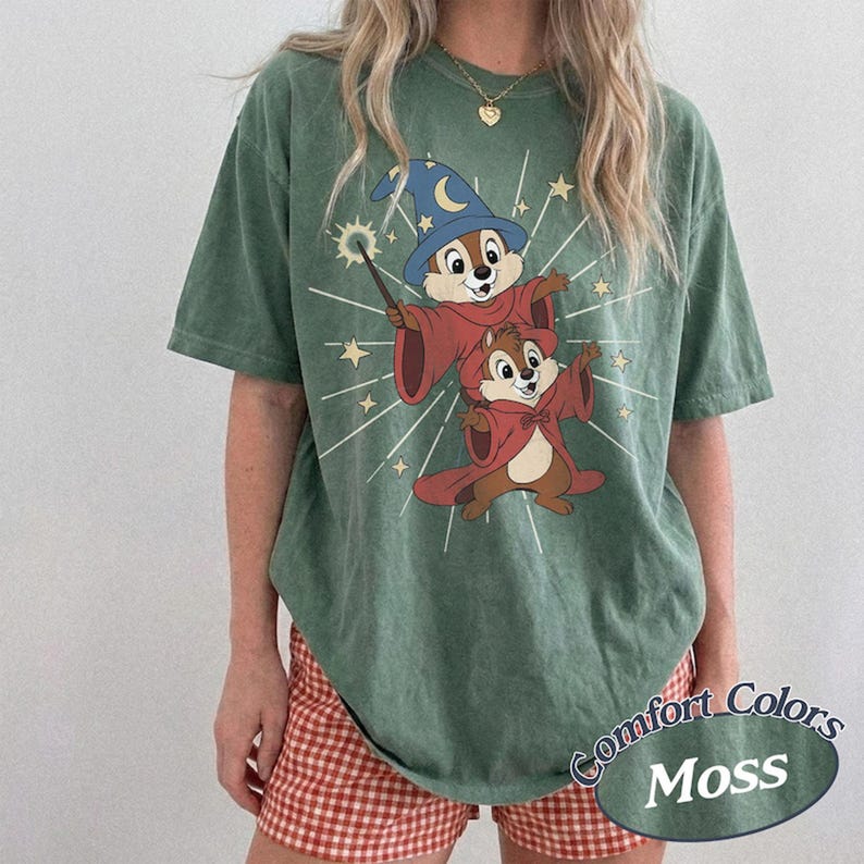 Op de afbeelding: Groen Comfort Colors T-shirt met een cartoon van twee eekhoorns als tovenaars, gekleed in rode gewaden en hoeden. Het shirt heeft de tekst "Comfort Colors Moss" in een cirkelvormig ontwerp.