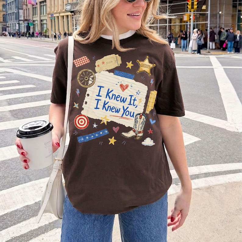 Puede incluir: Una camiseta de manga corta de color marr&oacute;n oscuro con un gr&aacute;fico central de papel de cuaderno rayado con el texto "I Knew It, I Knew You" en azul. El dise&ntilde;o incluye una bota de vaquero, una estrella de sheriff, una rueda y estrellas.