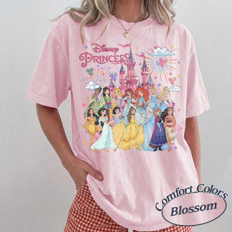 May include: Pink Comfort Colors t-shirt featuring a graphic of Disney Princesses in front of a castle with fireworks. The text "Disney Princess" is above the image. The shirt has short sleeves and a round neckline.