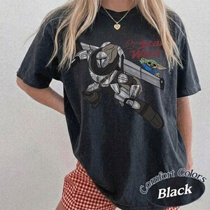 Pu&ograve; includere: T-shirt nera con grafica di Star Wars con The Mandalorian e Grogu. Il design include il testo "Star Wars" in rosso e le parole "Comfort Colors Black" in un design circolare.