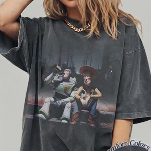 May include: A charcoal gray oversized t-shirt featuring a graphic print of Buzz Lightyear and Woody sitting together. The shirt has a vintage-washed appearance and includes the text Comfort Colors Pepper printed in the bottom right corner.