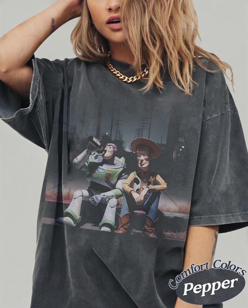 May include: A charcoal gray oversized t-shirt featuring a graphic print of Buzz Lightyear and Woody sitting together. The shirt has a vintage-washed appearance and includes the text Comfort Colors Pepper printed in the bottom right corner.