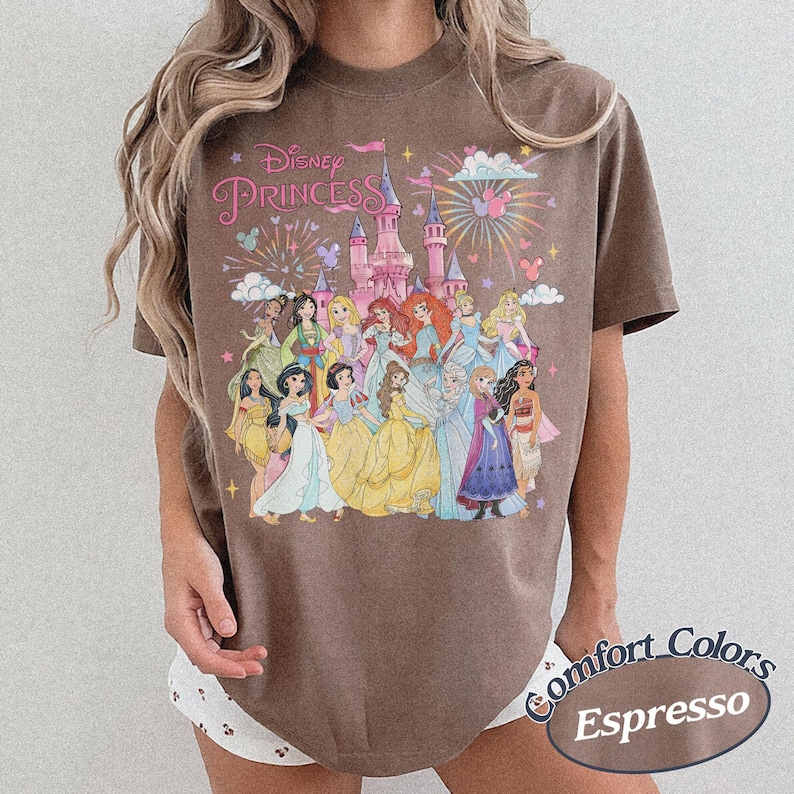 May include: Espresso-colored t-shirt featuring a Disney Princess graphic. The design includes a castle, fireworks, and various princess characters. The words "Disney Princess" are arched across the top. The shirt is part of the Comfort Colors collection.