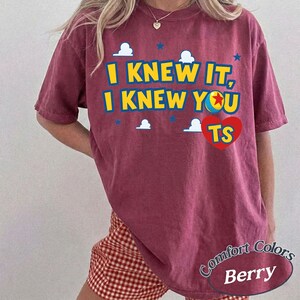 K&ouml;nnte beinhalten: Ein beerenfarbenes Kurzarm-T-Shirt mit dem gelben Text I KNEW IT, I KNEW YOU, einer bunten Ballgrafik und einem roten Herz mit den Buchstaben TS. Das Design enth&auml;lt kleine wei&szlig;e Wolken und blaue Sterne als Akzente auf der Vorderseite.