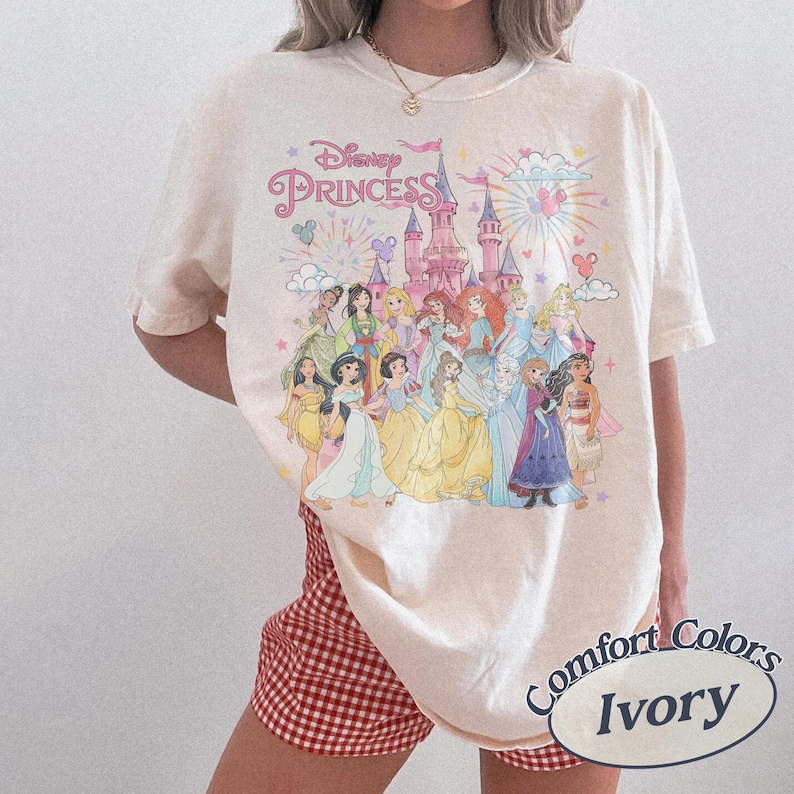 May include: Ivory-colored t-shirt featuring a Disney Princess graphic. The design includes the text "Disney Princess" above an illustration of several princesses in front of a castle. The shirt is a Comfort Colors brand.
