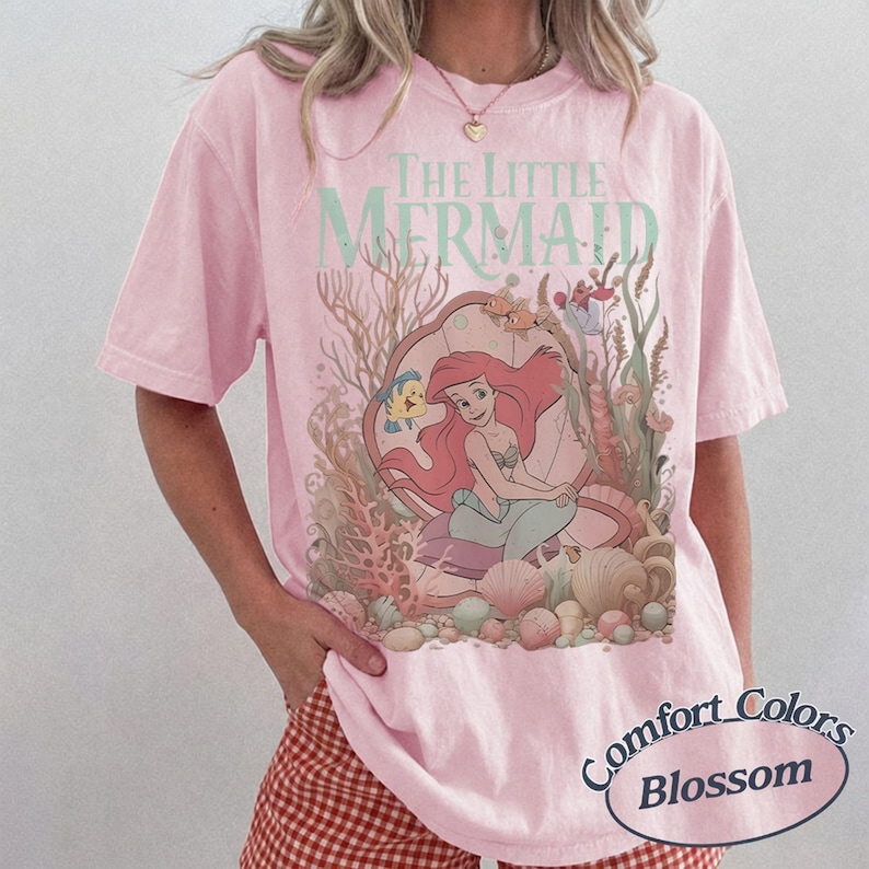Pu&ograve; includere: Una t-shirt a maniche corte rosa chiaro con una stampa grafica de La Sirenetta che raffigura Ariel seduta in una conchiglia circondata da coralli e vita marina. Il testo The Little Mermaid &egrave; stampato in verde chiaro sopra l'illustrazione.