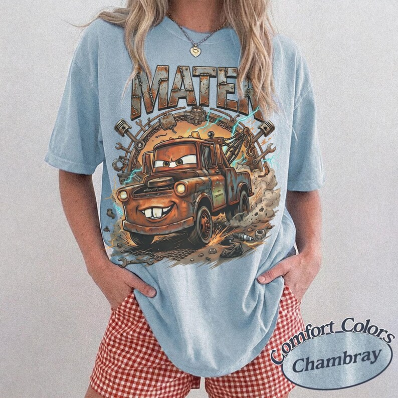 May include: A light blue short-sleeve t-shirt featuring a graphic print of a rusty brown tow truck character with the text MATER above it. The design includes mechanical parts like gears and pistons surrounding the truck with blue lightning.