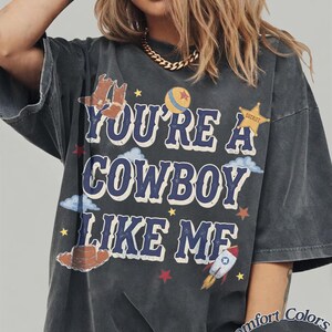 Op de afbeelding: Een antracietgrijs T-shirt met korte mouwen en de tekst "YOU'RE A COWBOY LIKE ME" in het blauw met witte omlijning. De afbeelding bevat illustraties van cowboylaarzen, een bal, een sheriffster, wolken, een hoed en een raket.