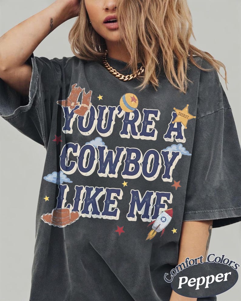 Op de afbeelding: Een antracietgrijs T-shirt met korte mouwen en de tekst "YOU'RE A COWBOY LIKE ME" in het blauw met witte omlijning. De afbeelding bevat illustraties van cowboylaarzen, een bal, een sheriffster, wolken, een hoed en een raket.