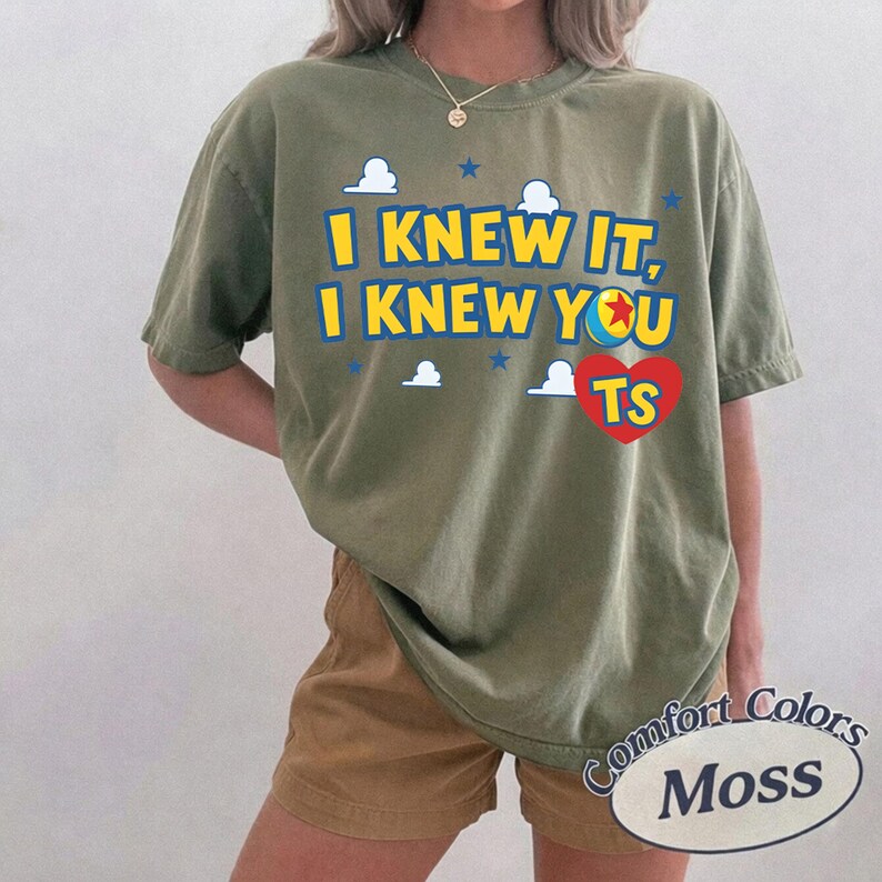 K&ouml;nnte beinhalten: Ein olivgr&uuml;nes Kurzarm-T-Shirt mit dem gelben Schriftzug I KNEW IT, I KNEW YOU TS, einem roten Herz und einer bunten Ballgrafik. Das Design enth&auml;lt kleine wei&szlig;e Wolken und blaue Sterne, die um den Text auf der Vorderseite des Shirts verteilt sind.