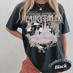 Może przedstawiać: Czarny t-shirt z kr&oacute;tkim rękawem z nadrukiem graficznym przedstawiającym zamek, motyle i kwiaty. Tekst brzmi Today was a FAIRY TALE oraz Can you feel the magic in the air? Wz&oacute;r zawiera delikatne odcienie r&oacute;żu, błękitu i złota na ciemnej tkaninie.