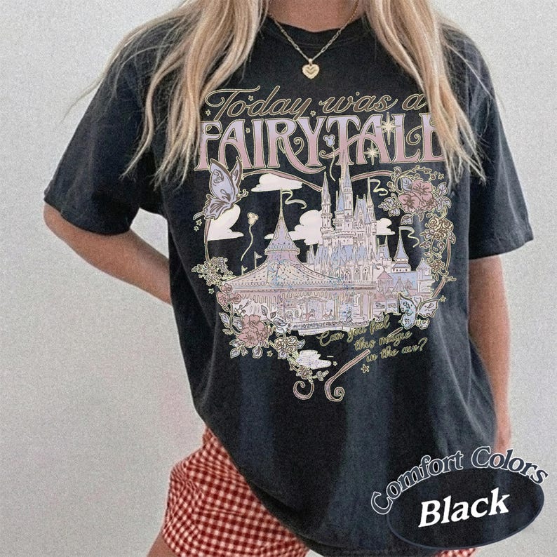 Może przedstawiać: Czarny t-shirt z kr&oacute;tkim rękawem z nadrukiem graficznym przedstawiającym zamek, motyle i kwiaty. Tekst brzmi Today was a FAIRY TALE oraz Can you feel the magic in the air? Wz&oacute;r zawiera delikatne odcienie r&oacute;żu, błękitu i złota na ciemnej tkaninie.