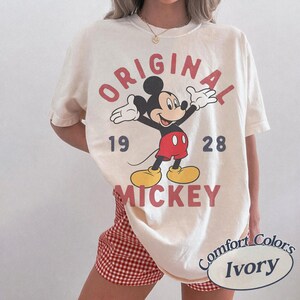 Op de afbeelding: Een ivoorkleurig T-shirt met korte mouwen en een afbeelding van Mickey Mouse met gespreide armen. De tekst ORIGINAL staat boven het personage en MICKEY eronder, met de cijfers 19 en 28 aan weerszijden. De afbeelding gebruikt rood, zwart en geel.