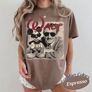 May include: Espresso-colored t-shirt featuring a vintage graphic of Mickey Mouse and Walt Disney. The word "Walt" is in red glitter script above the image. The shirt is a Comfort Colors brand.
