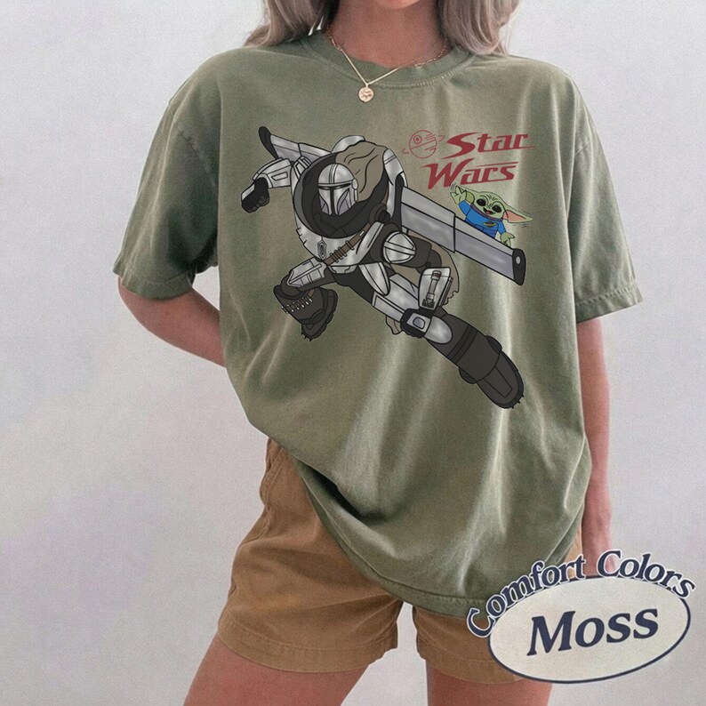 Pu&ograve; includere: T-shirt verde oliva con una grafica di un personaggio di Star Wars e Grogu. Le parole "Star Wars" sono stampate in rosso. La maglietta &egrave; etichettata "Comfort Colors Moss".