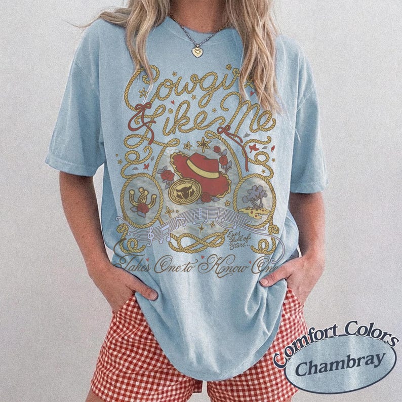 Op de afbeelding: Een lichtblauw t-shirt met korte mouwen en een grafische print met de tekst Cowgirl Like Me en Eyes full of Stars... Takes One to Know One. Het ontwerp bevat een rode cowboyhoed, een hoefijzer en touwelementen in een beige kleur.