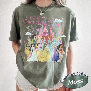 May include: Moss green t-shirt featuring a Disney Princess graphic. The design includes a pink castle, fireworks, and illustrations of various Disney princesses. The shirt has the words "Disney Princess" in pink script.