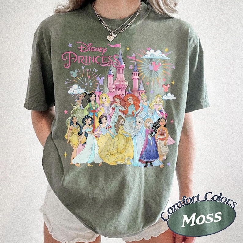 May include: Moss green t-shirt featuring a Disney Princess graphic. The design includes a pink castle, fireworks, and illustrations of various Disney princesses. The shirt has the words "Disney Princess" in pink script.
