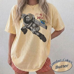 Pu&ograve; includere: T-shirt color burro con grafica di un personaggio con un'arma e una piccola creatura verde. Il testo "S Wars" &egrave; sopra la grafica. La maglietta &egrave; etichettata "Comfort Colors Butter".