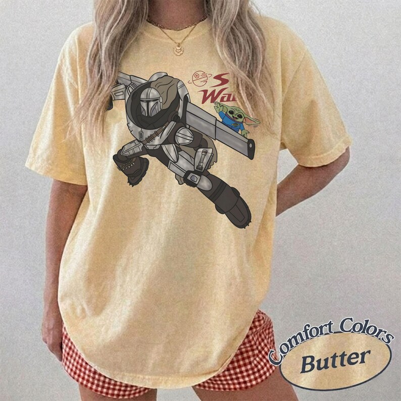 Pu&ograve; includere: T-shirt color burro con grafica di un personaggio con un'arma e una piccola creatura verde. Il testo "S Wars" &egrave; sopra la grafica. La maglietta &egrave; etichettata "Comfort Colors Butter".