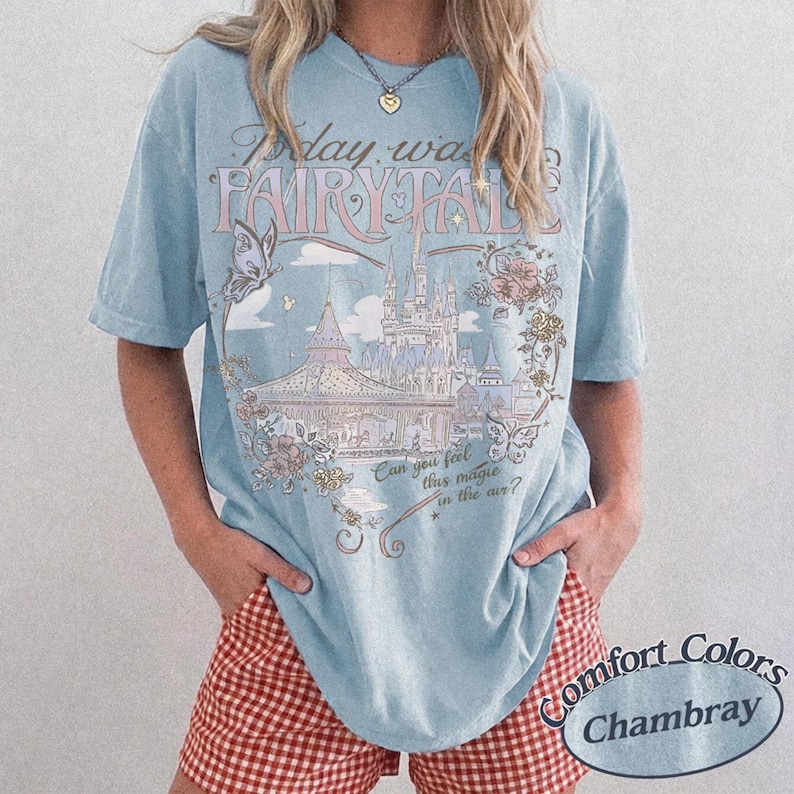 Może przedstawiać: Jasnoniebieski t-shirt z nadrukiem graficznym przedstawiającym zamek, motyle i elementy kwiatowe. Tekst głosi Today was a FAIRYTALE oraz Can you feel this magic in the air? Koszulka ma kr&oacute;tkie rękawy i okrągły dekolt o luźnym kroju.