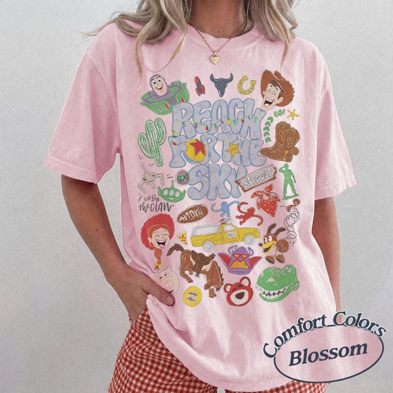 Peut inclure: Un t-shirt rose clair &agrave; manches courtes arborant une impression graphique color&eacute;e de divers personnages et objets de dessin anim&eacute; de la franchise Toy Story, avec le texte REACH FOR THE SKY en grandes lettres bleues au centre du v&ecirc;tement.