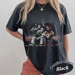 May include: A black short-sleeve t-shirt featuring a graphic print of the characters Buzz Lightyear and Woody sitting side-by-side. The shirt has a crew neckline and a relaxed fit. The graphic shows the two characters in a dark, outdoor setting.