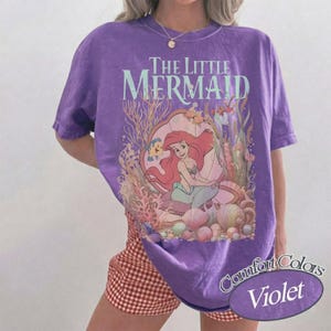 Pu&ograve; includere: Una t-shirt viola a maniche corte con una stampa grafica de La Sirenetta che raffigura Ariel seduta in una grande conchiglia circondata da coralli e vita marina. Il testo The Little Mermaid &egrave; stampato in un carattere azzurro sopra l'illustrazione.