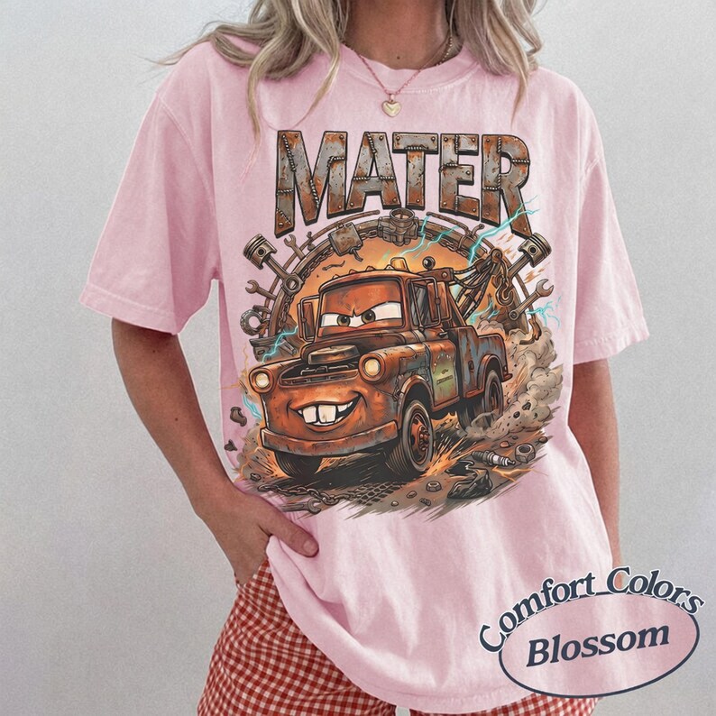 May include: A light pink short-sleeve t-shirt featuring a graphic print of a rusty brown tow truck character with large eyes and teeth. The text MATER is printed in a metallic, weathered font above the truck, surrounded by mechanical parts.