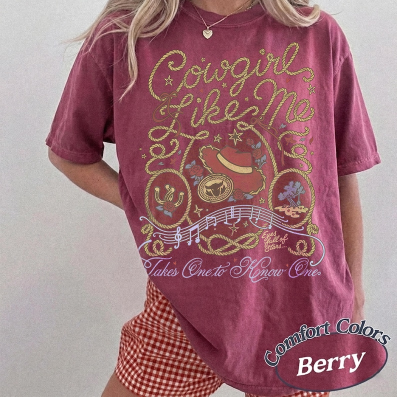 Op de afbeelding: Een bessenkleurig t-shirt met korte mouwen en een grafische print van een lasso, een cowboyhoed, een hoefijzer en muzieknoten. De tekst luidt Cowgirl Like Me, Takes One to Know One en de zin Eyes full of Stars is ook afgebeeld.