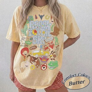 Peut inclure: Un t-shirt jaune beurre &agrave; manches courtes avec une impression graphique portant le texte Reach For The Sky et diverses illustrations de personnages de dessins anim&eacute;s, dont une cow-girl, un cochon, un dinosaure, un camion jaune et une botte.