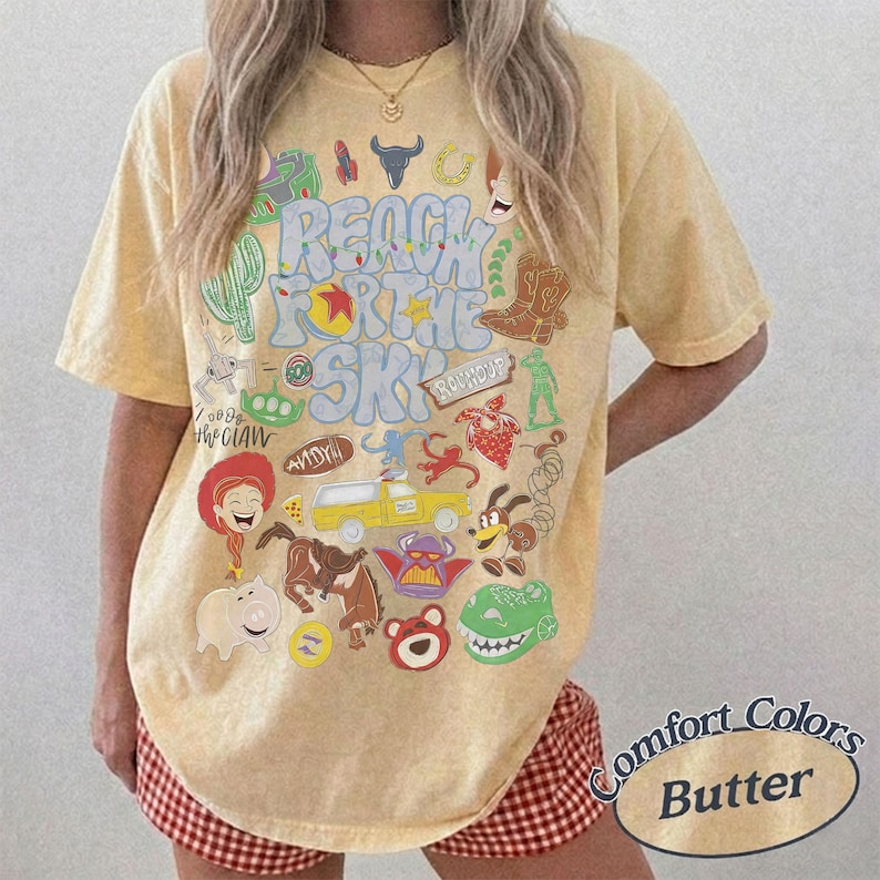 Peut inclure: Un t-shirt jaune beurre &agrave; manches courtes avec une impression graphique portant le texte Reach For The Sky et diverses illustrations de personnages de dessins anim&eacute;s, dont une cow-girl, un cochon, un dinosaure, un camion jaune et une botte.
