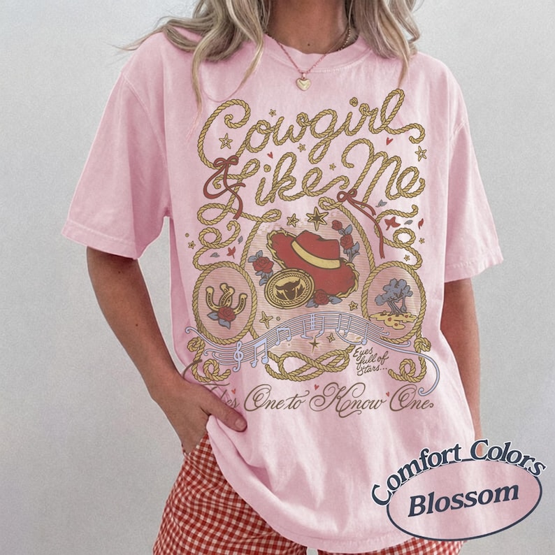 Op de afbeelding: Een lichtroze T-shirt met korte mouwen en een grafische print met de tekst Cowgirl like Me, Eyes full of Stars en One to Know One. Het ontwerp bevat illustraties van een rode cowboyhoed, een hoefijzer, een touwrand en muzieknoten.