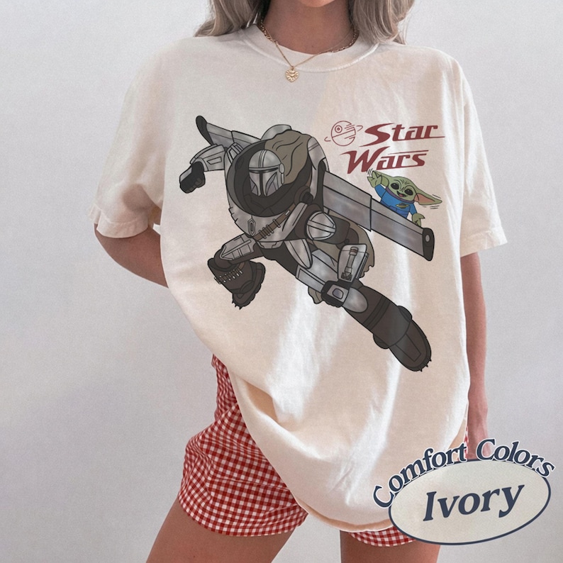 Pu&ograve; includere: T-shirt color avorio con una grafica di The Mandalorian e Grogu di Star Wars. Il design include il logo di Star Wars in rosso. La maglietta &egrave; etichettata "Comfort Colors" e "Ivory".