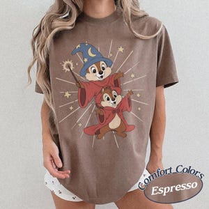 Op de afbeelding: Espresso-kleurig T-shirt met een cartoonillustratie van twee chipmunks verkleed als tovenaars, een met een blauwe hoed en toverstaf. Het ontwerp bevat sterren en stralende lijnen. De tekst "Comfort Colors Espresso" staat rechtsonder.