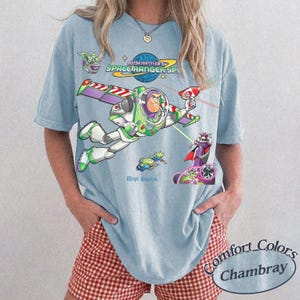 May include: Light blue t-shirt featuring a graphic of Buzz Lightyear in flight, with the text "Buzz Lightyear Space Ranger SP" and "Magic Kingdom." The shirt also has the Comfort Colors Chambray logo.