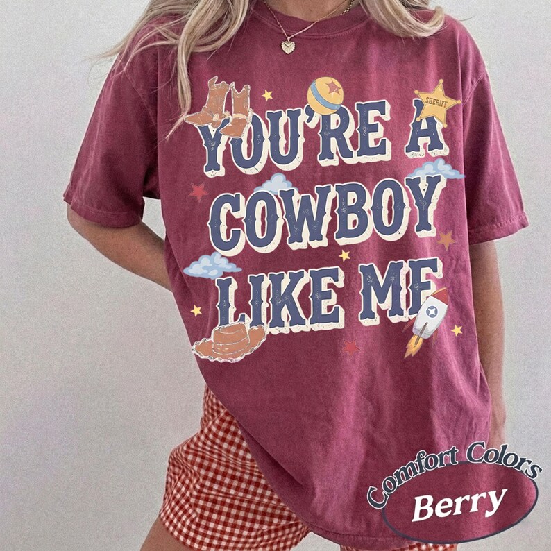 Op de afbeelding: Een bessenkleurig T-shirt met korte mouwen en de tekst "YOU'RE A COWBOY LIKE ME" in blauwe letters. De afbeelding bevat illustraties van cowboylaarzen, een sheriffster, een bal, een hoed, wolken, sterren en een raket in diverse kleuren.